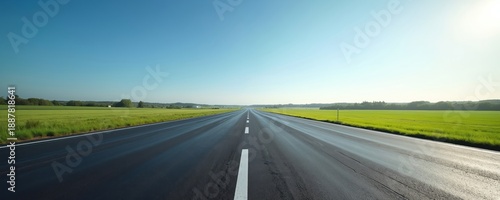 Empty asphalt road stretches to horizon through green fields under clear blue sky. Bright sun shines on the smooth highway. Countryside landscape offers endless journey and peaceful drive.