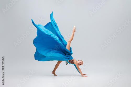ballerina performing an acrobatic move stands on her hands with a leg raised high. Cheerleader is wearing a sport leotard and a flowing blue cloth, light gray background accentuates dynamic motion.