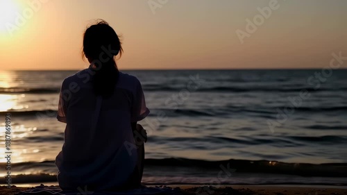 Silhouetted figure of a woman sitting on the beach at sunset, gazing out at the calm sea, serene atmosphere