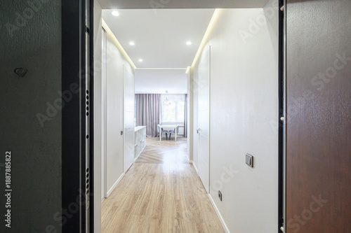 Modern hallway with light wood flooring, white walls, recessed ceiling lights, LED strip lighting, sliding doors, leading to dining area with table and curtains.