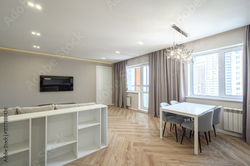 Modern living room with beige sofa, white shelving, flat-screen TV, dining area with white table and grey chairs, herringbone wood flooring, large windows with grey curtains, chandelier.