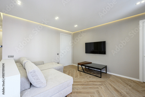 Minimalist living room with light grey sofa, herringbone wooden floor, wall-mounted TV, sleek black coffee table, recessed ceiling lights, neutral walls, clean lines.