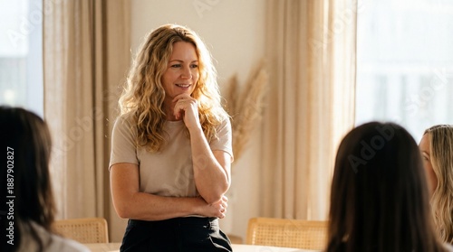 Confident woman with wavy blonde hair smiling and speaking to small group in bright modern meeting room, warm natural light and neutral decor creating relaxed atmosphere