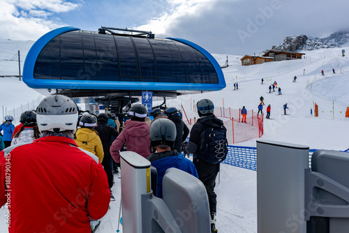 Photos Skiers in helmets queue at a modern chairlift station, passing through turnstiles while others descend groomed snowy slopes near alpine buildings and fencing