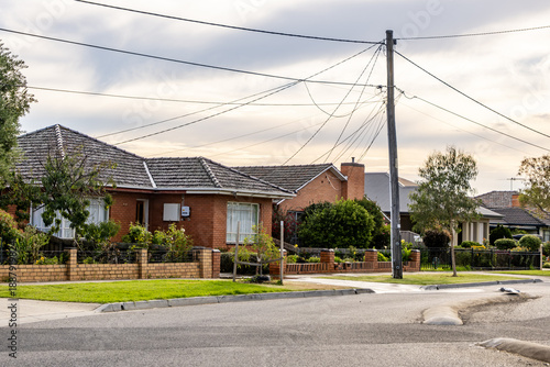 Fotografía A quiet suburban residential street with single-storey brick houses, front gardens, and overhead power lines at a street corner
