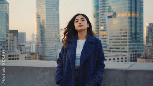 Cinematic video of young woman standing still on rooftop in warm coat during blue hour with city lights