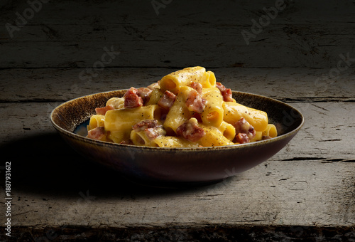 Dark bowl with rigatoni pasta carbonara on rough wood