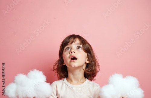 Little girl with brown hair looks up with amazement holding fluffy white clouds. Child has open mouth mouth in surprise against a pink backdrop. She is fascinated by something in the sky above.