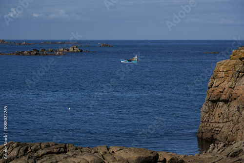 A fishing boat sails on the blue sea, surrounded by rocky islands and cliffs, showcasing the serene beauty of Brittany. Plage des Amiets, France.