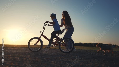 A boy rides a bike while a woman helps him on the dirt path. The boy moves the bike forward as the woman supports him under the setting sun.