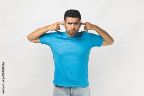 Portrait of annoyed sad unshaven man wearing blue T- shirt standing covering ears with fingers, does't want to hear unpleasant sounds. Indoor studio shot isolated on gray background.
