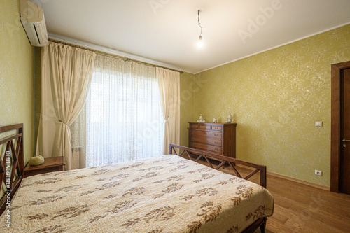 Wallpaper Mural A cozy bedroom with a wooden bed, dresser, and nightstand. Light green floral wallpaper, beige curtains, wooden flooring, and an air conditioner. Soft, traditional decor. Torontodigital.ca