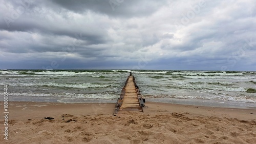An old wooden pier reaches into a dark sea where white waves break with force. Heavy clouds hang above, briefly revealing blue sky, capturing tension, movement and raw coastal atmosphere.
