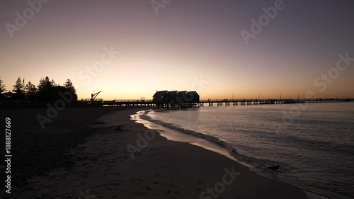 The famous mile-long jetty in Western Australia stretches into the ocean during golden hour as gentle waves crash along the shoreline. Warm sunset light, calm coastal colors, and rhythmic surf create 