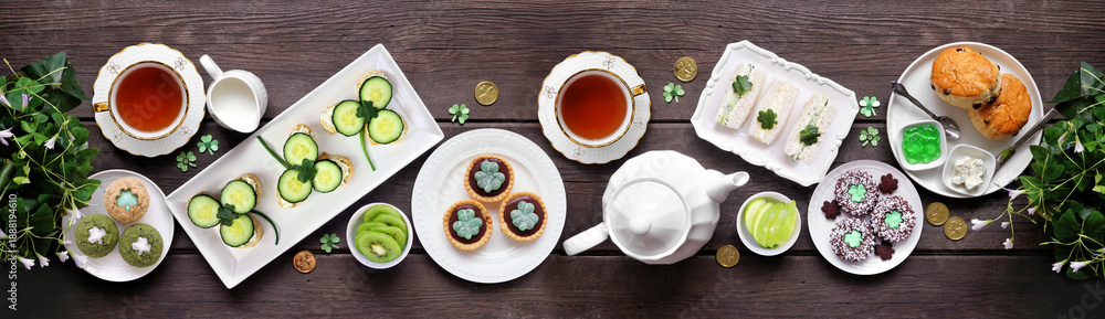 Fototapeta premium St Patrick's Day tea party table scene against a dark wood banner background. Variety of green shamrock theme finger sandwiches, fruits, scones and sweet desserts. Above view.
