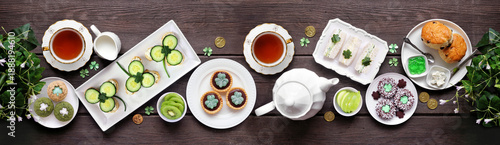 St Patrick's Day tea party table scene against a dark wood banner background. Variety of green shamrock theme finger sandwiches, fruits, scones and sweet desserts. Above view.
