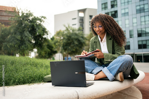 Young African American female student using laptop learning sitting in university garden park, studying online digital business course