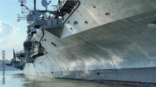 The radar mast and superstructure of the USS Intrepid rise above the deck in New York harbor. Antennas, satellite dishes, and steel structures show the ship's naval technology against a blue sky.