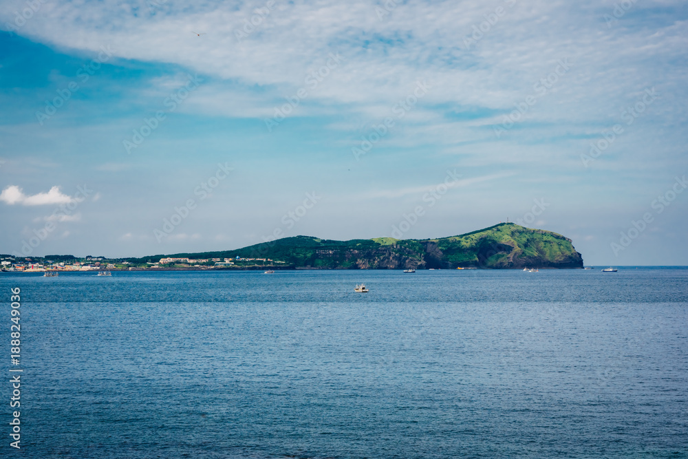 Naklejka premium A wide coastal seascape on Jeju Island, South Korea, with calm blue water and a green volcanic island on the horizon. Small boats dot the ocean under a bright sky with soft, textured clouds.