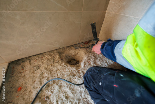 Billede på lærred A tradesperson in high visibility gear operates a rotary hammer, drilling a circular recess in a concrete subfloor beside beige tiles, with debris and cable on rough surface