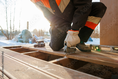 Quadro su tela A worker in insulated gear kneels fastening composite boards to a timber frame using a pneumatic nailer, with a spirit level and cordless drill nearby in winter light