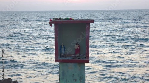 Dilapidated seaside monument with statues of Jesus Christ and Mother Mary placed on the shore, set against the ocean in a weathered coastal environment.