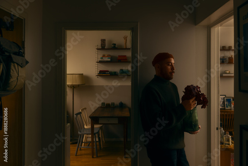 Slika na platnu A calm bearded man holding a vase of flowers stands in a cozy, softly lit apartment interior