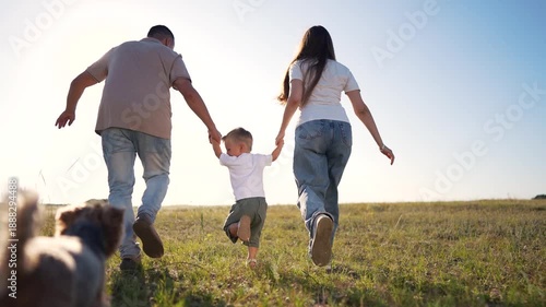 A man, woman, and boy run hand-in-hand through a tall grassy field. The boy skips as the man and woman hold his hands in the sunny grassy field under the blue sky.