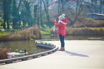 A young girl in a warm winter outfit taking a photo of a duck on a wooden pathway in a park near the water.