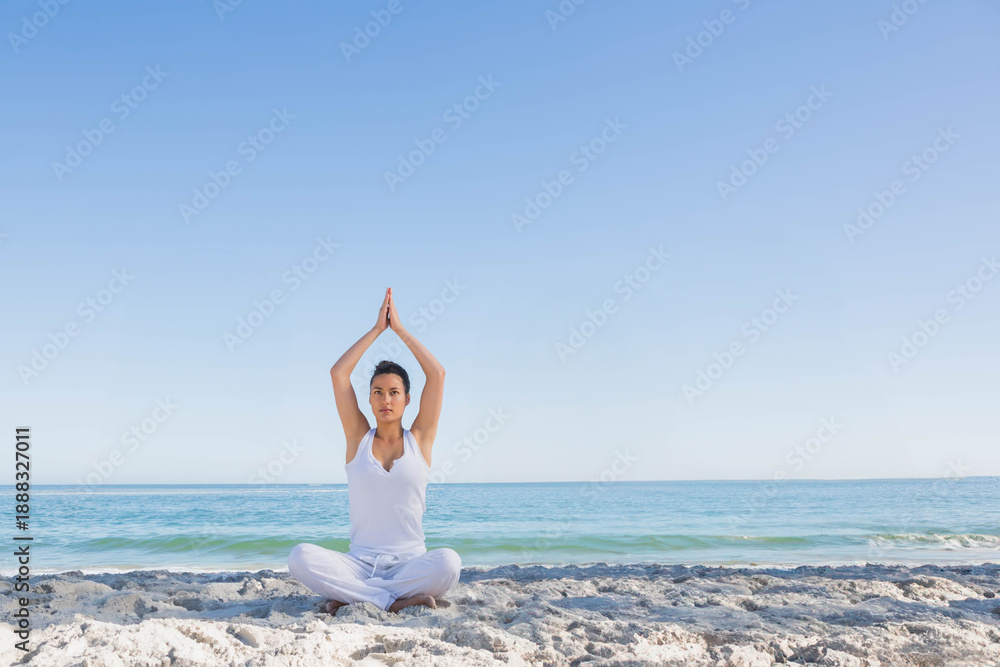 Fototapeta premium Woman practicing yoga and meditating while sitting cross legged on sand near ocean in white