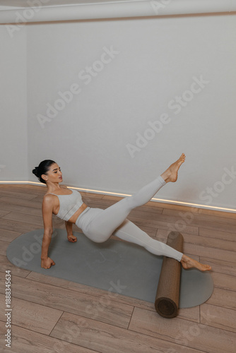 Pilates foam roller. Athletic brunette woman practices pilates on foam roller wearing light grey outfit on grey mat. Controlled exercises show strength flexibility balance and body control.