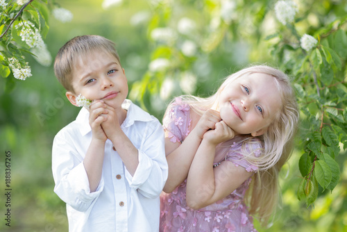 A little boy and a little girl are posing in a blooming garden and smiling at the camera.