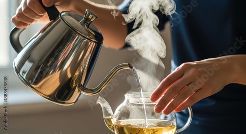 Person pouring hot tea from a silver kettle into a clear glass teapot with steam rising in a cozy