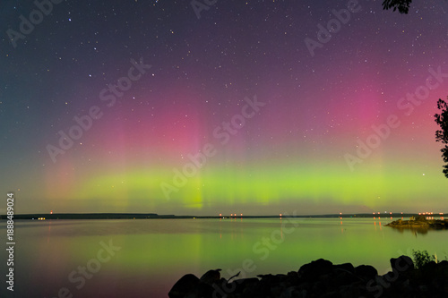 Aurora borealis illuminated over a lake in the night sky painting vibrant ribbons of light. Green, purple, and pink aurora pillars of light dance across the northern horizon creating a magical display