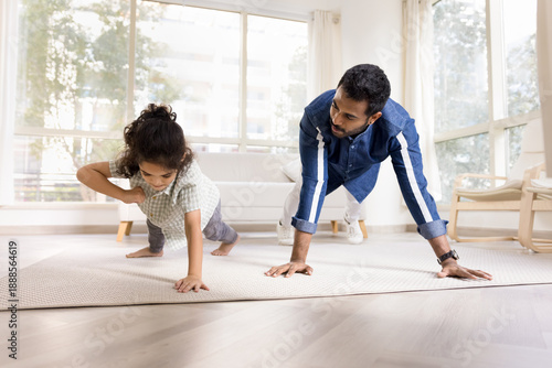 Young Indian man and his little 6s little kid boy engaging in sports activity in spacious, modern living room, performing push-up exercises on floor. Father-son bonding, family fitness, health-care
