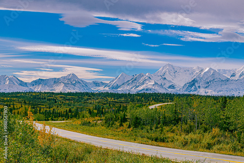 Fotografía Scenic view of a winding highway cutting through Alaska’s tundra toward the snow-covered peaks of the Alaska Range under a dramatic blue sky, photographed between Fairbanks and Valdez