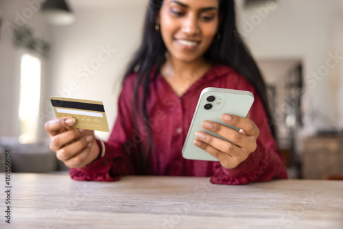 Positive young Indian consumer woman paying for purchases by credit card, using mobile phone for shopping on e-commerce platform, managing account on bank financial application. Cropped close up shot