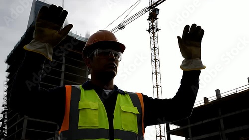 Construction worker wearing safety gear and protective gloves gives hand signals at a busy outdoor job site near tall structures
