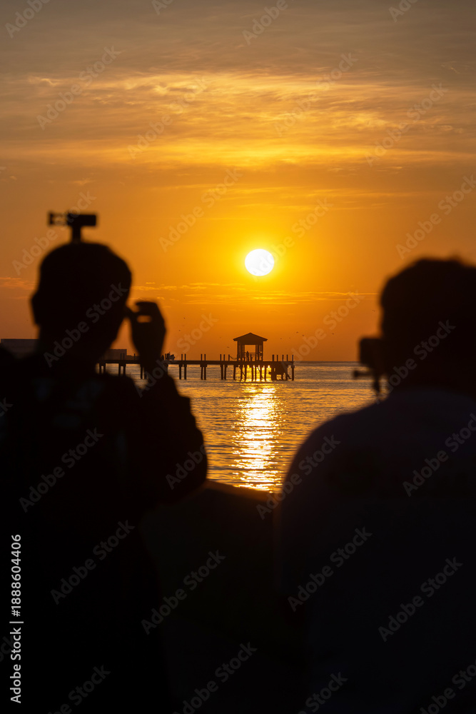 Fototapeta premium Silhouetted photographers capturing golden sunset over pier and calm sea at Bang Pu in Samut Prakan, Thailand. Popular spot to relax, enjoy sunrise and sunsets.