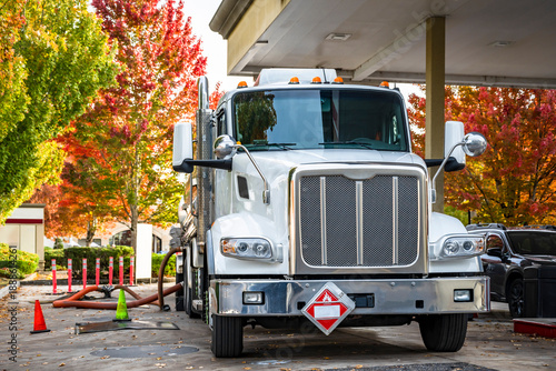 Day cab white big rig fuel semi truck truck with a tanker semi trailer unloads delivered fuel at gas station on city street with autumn trees