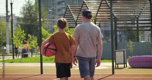 Father walks with son on outdoor basketball court during summer day. Father supports child before sport activity. Trust and care. Dad represents healthy lifestyle and modern parenting. Happy boy child