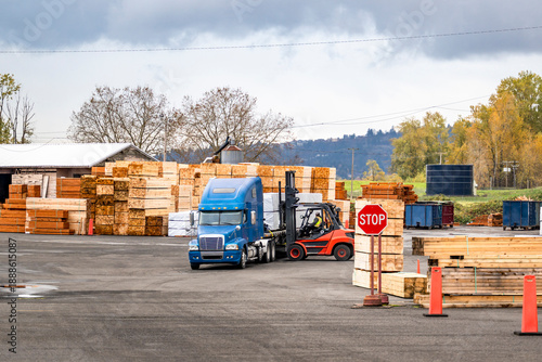 Fork lift loading lumber cargo on blue big rig semi truck with flat bed semi trailer standing on the warehouse parking lot