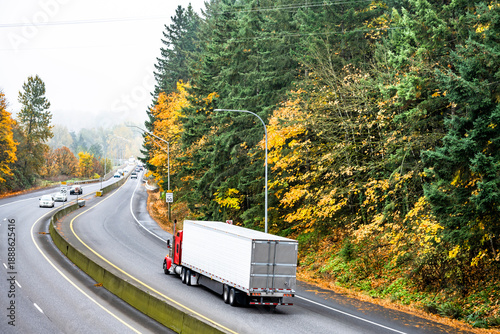 Red bonnet big rig semi truck transporting cargo in refrigerator semi trailer running on the autumn curved road with green and yellow trees and fog