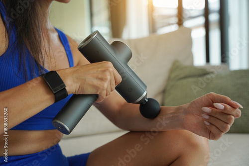 Woman using percussion massage gun post-workout recovery seated on sofa in bright living room