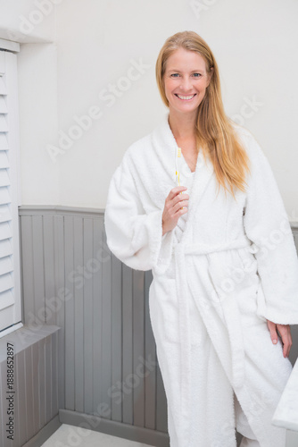 Mid-adult woman standing in small bathroom wearing white terry bathrobe holding toothbrush