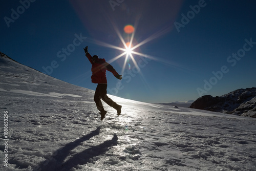 Person is jumping on sunlit snowy slope, wearing red jacket and casting shadow with lens flare