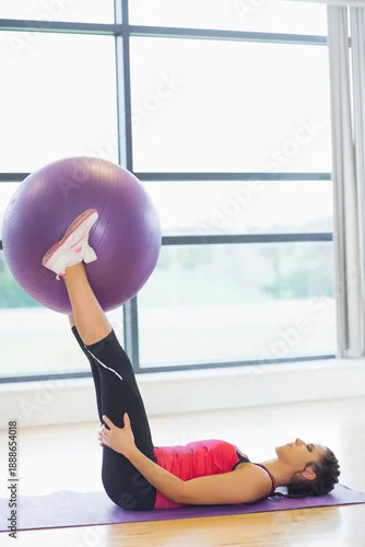 Fototapeta Woman is lying on purple mat near large windows on wood floor and is holding ball