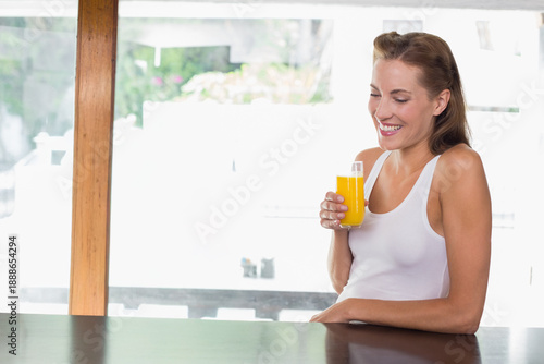 Woman in white tank sitting at counter by window, holding glass of orange juice, copy space