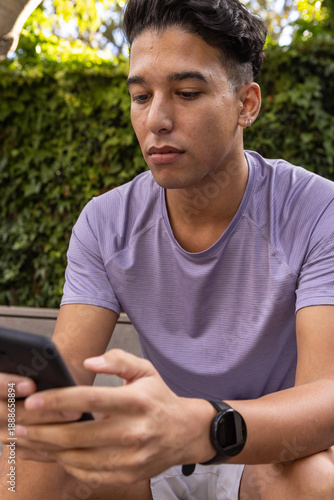 Obraz na plátně Man in early 20s sitting on garden bench wearing sportswear, checking smartphone