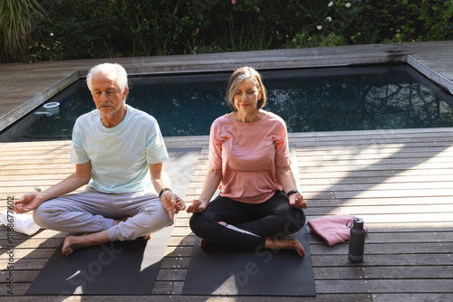 Senior couple wearing activewear meditating on black yoga mats on poolside deck with water bottle